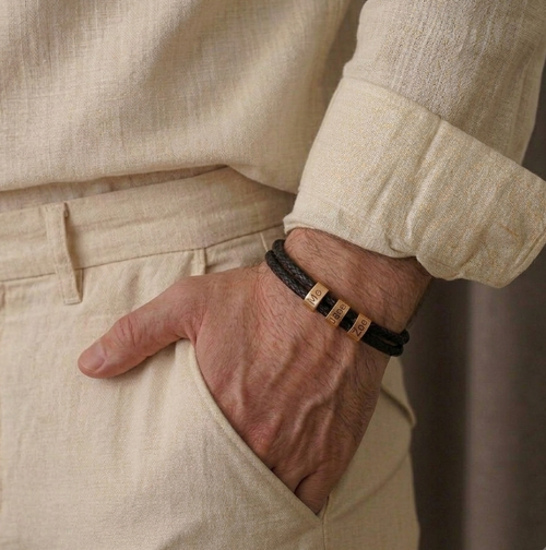 A close-up of a person's hand in a pocket wearing a black leather bracelet with three gold-tone metal accent cubes engraved with 'MADE IN ITALY'. The person is wearing a matching beige linen shirt and trousers.